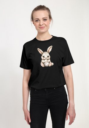 Kvinde står og smiler og har en sort T-shirt på med en sød illustreret kanin, der holder et plettet æg, trykt på brystet.