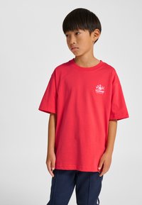 Röd bomulls-T-shirt med korta ärmar och rund halsringning. Har en vit logotyp på vänster bröst. Kombineras med marinblå byxor.