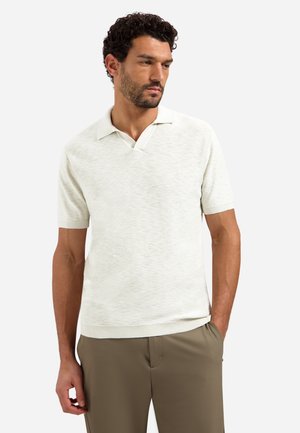 Man met kort krullend haar die een crèmekleurig gebreid poloshirt met korte mouwen en olijfgroene broek draagt, zijwaarts kijkt met één hand in zijn zak.