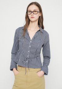 Γυναίκα με μακριά μαλλιά που φοράει γυαλιά με σκούρο σκελετό, μπλε καρό πουκάμισο με κουμπιά δεμένο στη μέση και μπεζ παντελόνι, με τα χέρια στις τσέπες, ουδέτερο φόντο.