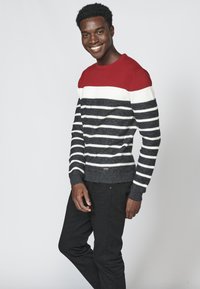 Maglione lavorato a maglia con un motivo a righe orizzontali rosse, bianche e nere. Maniche e fondo a costine, con dettaglio del logo all'altezza dei fianchi.