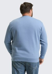 Man met kort donker haar, gekleed in een lichtblauwe sweatshirt en blauwe jeans, met zijn rug naar de camera toe tegen een effen lichte achtergrond.