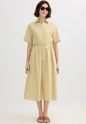 Mujer con vestido camisero midi beige de manga corta, sandalias marrones y gafas de sol oscuras, de pie contra un fondo liso y claro.