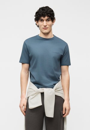 Moški kodrastih las, oblečen v modro majico s kratkimi rokavi in temne hlače, z bež jopico, vezano okoli pasu, stoji pred enobarvnim ozadjem.