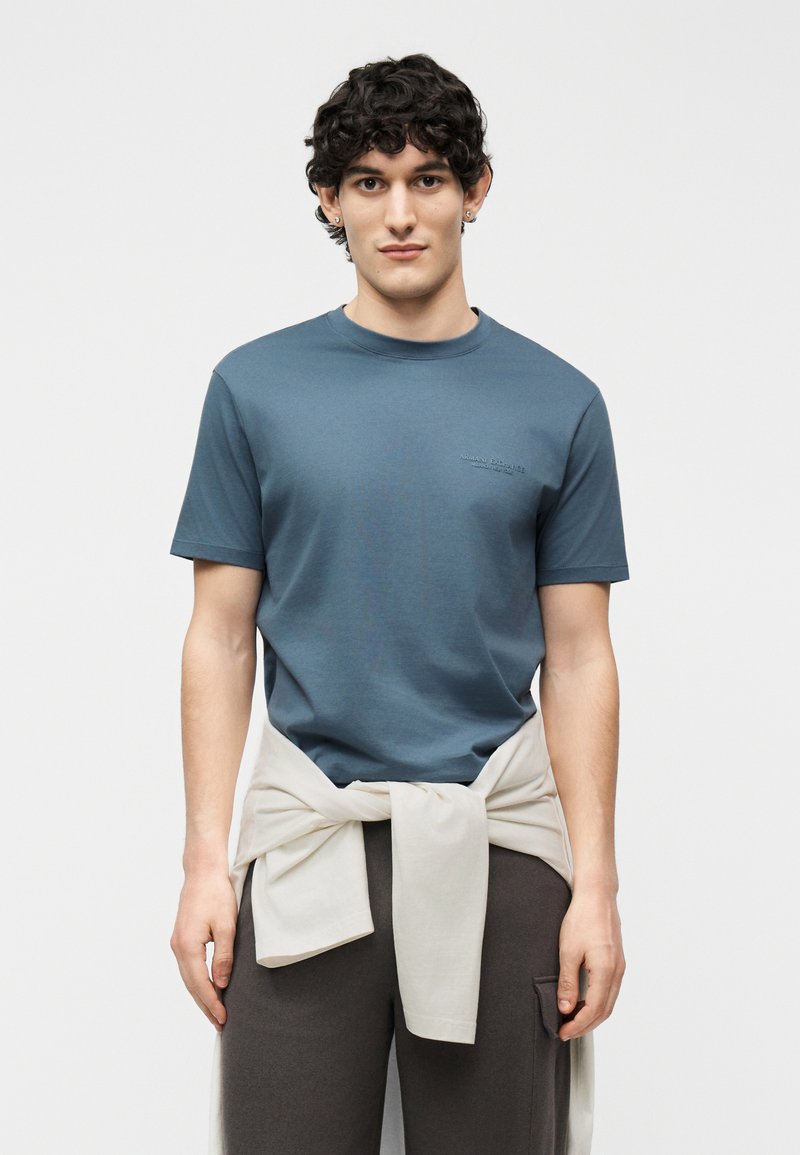 Άντρας με σγουρά μαλλιά, που φοράει μπλε μπλουζάκι και σκουρόχρωμο παντελόνι, με μπεζ φούτερ δεμένο γύρω από τη μέση του, στέκεται μπροστά σε ένα απλό φόντο.