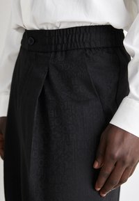 Black trousers with an elastic waistband, featuring a textured pattern and button detail. The fabric has a subtle sheen.