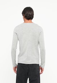 Pull-over en tricot gris à manches longues et col rond. Texture douce, silhouette ajustée et finition légèrement chinée. Jeans noirs visibles en dessous.