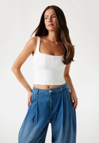 Bílý fitted crop top se čtvercovým výstřihem a texturovanými panely, kombinovaný s volnými modrými kalhotami s vysokým pasem, které mají sklady a knoflík.