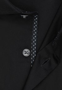 Tissu noir avec un bouton brillant, présentant une doublure intérieure à motifs noir et blanc. Le design comprend un col à revers et une ouverture de poche visible.