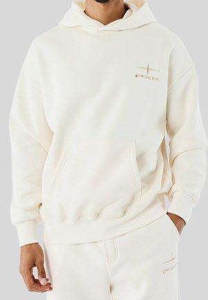 Mann trägt einen cremefarbenen Hoodie und passend dazu Sweatpants mit dezent besticktem Logo auf Brust und Oberschenkel, steht mit einer Hand in der Tasche.