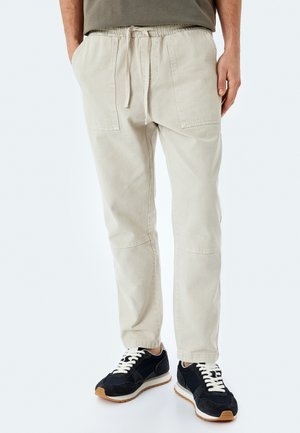 Pantalones jogger de algodón color beige con cintura elástica y cordón, que cuentan con dos bolsillos laterales y un diseño de pierna ajustada. Llevados con zapatillas negras.