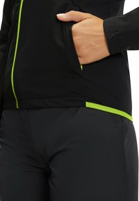 Zwarte lichtgewicht jas met een getailleerd ontwerp, voorzien van een groene rits en afwerkingen, gecombineerd met gladde zwarte broek. Zak zichtbaar op de jas.