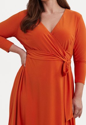 Vrouw draagt een lange mouw oranje wikkeljurk met een gestrikte taille en een V-hals, hand rustend op haar heup.
