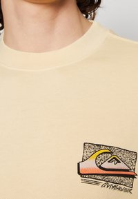 Sudadera de color crema con un logo gráfico en negro, amarillo y rosa. El cuello es acanalado, con una tela suave y texturizada.