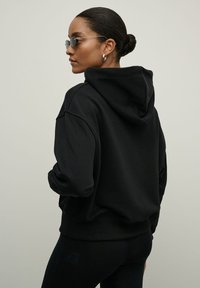 Juodas gobtuvu džemperis su laisvu siluetu, minkšta tekstūra ir ripsuota apykakle. Dėvimas su juodomis leginsais ir ovaliais akinius nuo saulės, suteikiantis elegantišką išvaizdą.