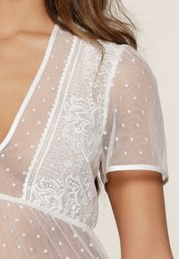 Transparante witte top met stippenpatroon, kantdetails, V-hals, korte mouwen en witte afwerking langs de halslijn en mouwen.
