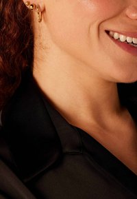 Des boucles d'oreilles en or et des clous d'oreilles adornent l'oreille. Le mannequin porte une chemise en satin noir, mettant en valeur un tissu lisse et brillant contre la peau.