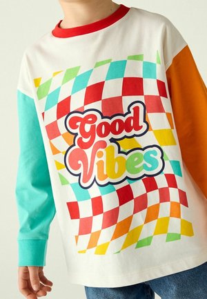 Enfant portant une chemise à manches longues colorée avec un col rouge, des manches turquoise et orange, et le texte "Good Vibes" sur un fond à carreaux vif.