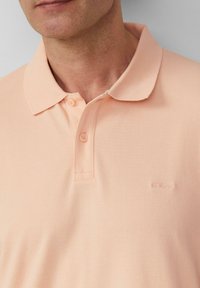 Hellpfirsichfarbendes Polohemd aus strukturiertem Stoff, mit Kragen und zwei Knöpfen am Hals. Verfügt über ein dezentes s.Oliver-Logo.