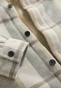 Karierte Bluse in Beige, Grau und Creme. Verfügt über schwarze Knöpfe und ein weiches, strukturiertes Innenfutter. Klassischer Kragen und Knopfmanschetten.