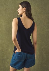 Sügavsinine V-kaelusega tank top, mis on valmistatud pehmest kangast, koos denim lühikesed püksid, millel on taskud ja vabalt langev lõige. Taust on tekstuuriga roheline.