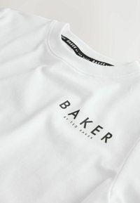 Balti crewneck marškinėliai su juodu užrašu ant krūtinės „BAKER BY TED BAKER“, rodoma arti viršutinės priekinės dalies ir apykaklės.