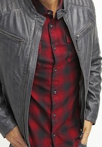 Grå læderjakke med quiltede skuldre og lynlåslommer, lagdelt over en rød og sort ternet skjorte med knaplukning.