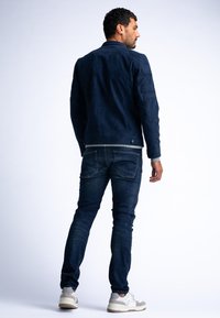 Giubbotto in suede blu navy con colletto classico, dotato di chiusura a bottoni e tasche laterali, abbinato a jeans scuri e sneakers grigie.
