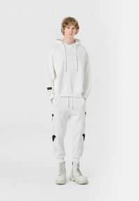 Sudadera blanca con bolsillo delantero y cordones, combinada con joggers blancos con patrones de hojas negras, usados con botas pesadas blancas.