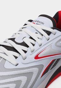 Κοντινό πλάνο σε λευκό, μαύρο και κόκκινο αθλητικό παπούτσι Reebok που δείχνει την υφή στο πάνω μέρος, τα κορδόνια, το λογότυπο στη γλώσσα και τα κόκκινα διακοσμητικά στη πλευρά.