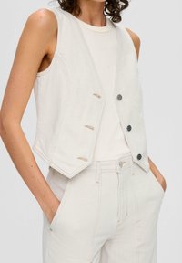 Beige Jeansweste mit taillierter Passform, tiefem V-Ausschnitt, vier Knöpfen und tonalen Nähten. Getragen über einem enganliegenden cremefarbenen Oberteil und passenden Hosen.