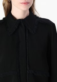 Sort bluse med krave og frynser, glat tekstur og knaplukning. Stoffet er let og semi-gennemsigtigt.