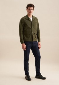 Olivgrüne Jacke mit langen Ärmeln, Stehkragen, zwei frontalen Taschen und Knopfverschluss, kombiniert mit blauen Hosen und schwarzen Schuhen.