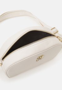 Bílá kožená crossbody taška s zaobleným tvarem, zlatým kováním a zipovým uzavřením, které odhaluje černý interiér. Součástí je nastavitelné rameno.