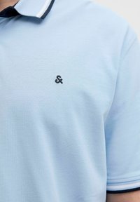 Gaiši zils polo krekls ar teksturētu audumu, ar melnu izšūtu logotipu un tumši zilu apkakli ar baltām svītrām.