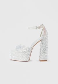 Srebrni platformni čevlji z okraski iz kristalčkov in odprtim prednjim delom. Imajo trak okoli gležnja in teksturiran blok peto.