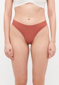 Tanga kupić u visokom struku u boji rđe, izrađena od glatkog, rastezljivog materijala. Ima minimalistički dizajn s spojenim bočnim šavovima.