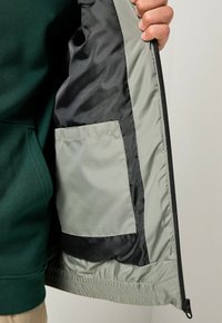 Leichte Jacke mit grünem Außenmaterial und grauem Innenfutter. Verfügt über ein schwarzes Satinfutter und eine kleine Tasche aus hellgrauem Stoff.