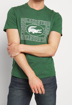 Homme portant un t-shirt vert Lacoste avec un logo crocodile blanc et le texte "Lacoste Classics Since 1927", les bras croisés, assorti à un jean foncé.