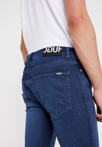 Mörkblå denimjeans med slim fit, med en svart etikett med vit text vid midjan och två bakfickor.