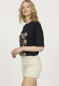 T-shirt en coton noir avec imprimé floral rouge et beige, associé à un short en denim beige. Le short présente des ourlets roulés et des poches arrière.