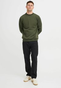 Oliven grønn genser med rund hals og ribbestrikkede mansjetter og nedkant, parret med svarte bukser og hvite sneakers med brune striper.