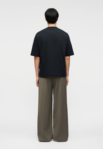 Άτομο που στέκεται με την πλάτη και φοράει μια φαρδιά μαύρη κοντομάνικη μπλούζα και φαρδιά καφέ παντελόνια μπροστά σε απλό φόντο.