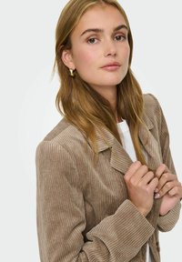 Beige kordflöjtsblazer med krage, synlig struktur från ribbningen och stora manschetter. Modellen håller i reversen och har på sig en vit topp.