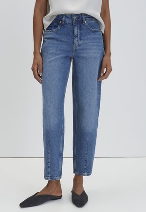 Mujer con vaqueros azules de talle alto y pierna recta, combinados con mules planos negros puntiagudos, sobre un fondo liso.