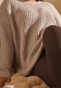 Person som bär en texturerad beige stickad tröja och mörkbruna leggings, sitter på en mjuk beige filt.