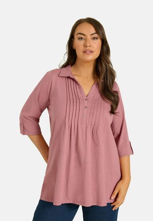 Vrouw met lang bruin haar die een stoffig roze blouse met driekwartmouwen, knoop- en plooi-details draagt, gecombineerd met donkerblauwe broek.
