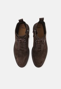 Botine din suede maroniu, cu vârful rotund, șireturi în față și detaliu cu fermoar pe lateral. Talpă plată din cauciuc și tab pentru strângere din material în spatele botinei.