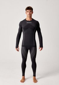 Zwarte lange mouwen compressietop en legging met gaaspanelen, reflecterende accenten en een opvallend logo op de borst. Strak aansluitende pasvorm.