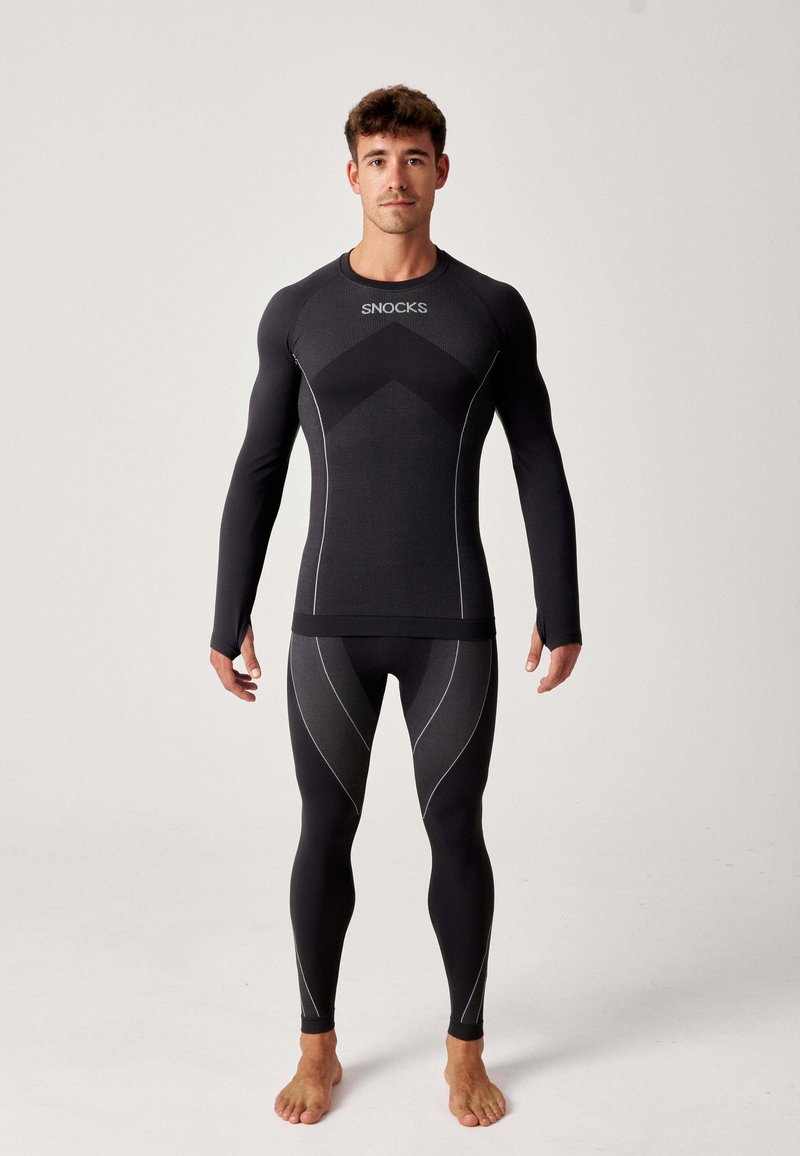 Zwarte lange mouwen compressietop en legging met gaaspanelen, reflecterende accenten en een opvallend logo op de borst. Strak aansluitende pasvorm.